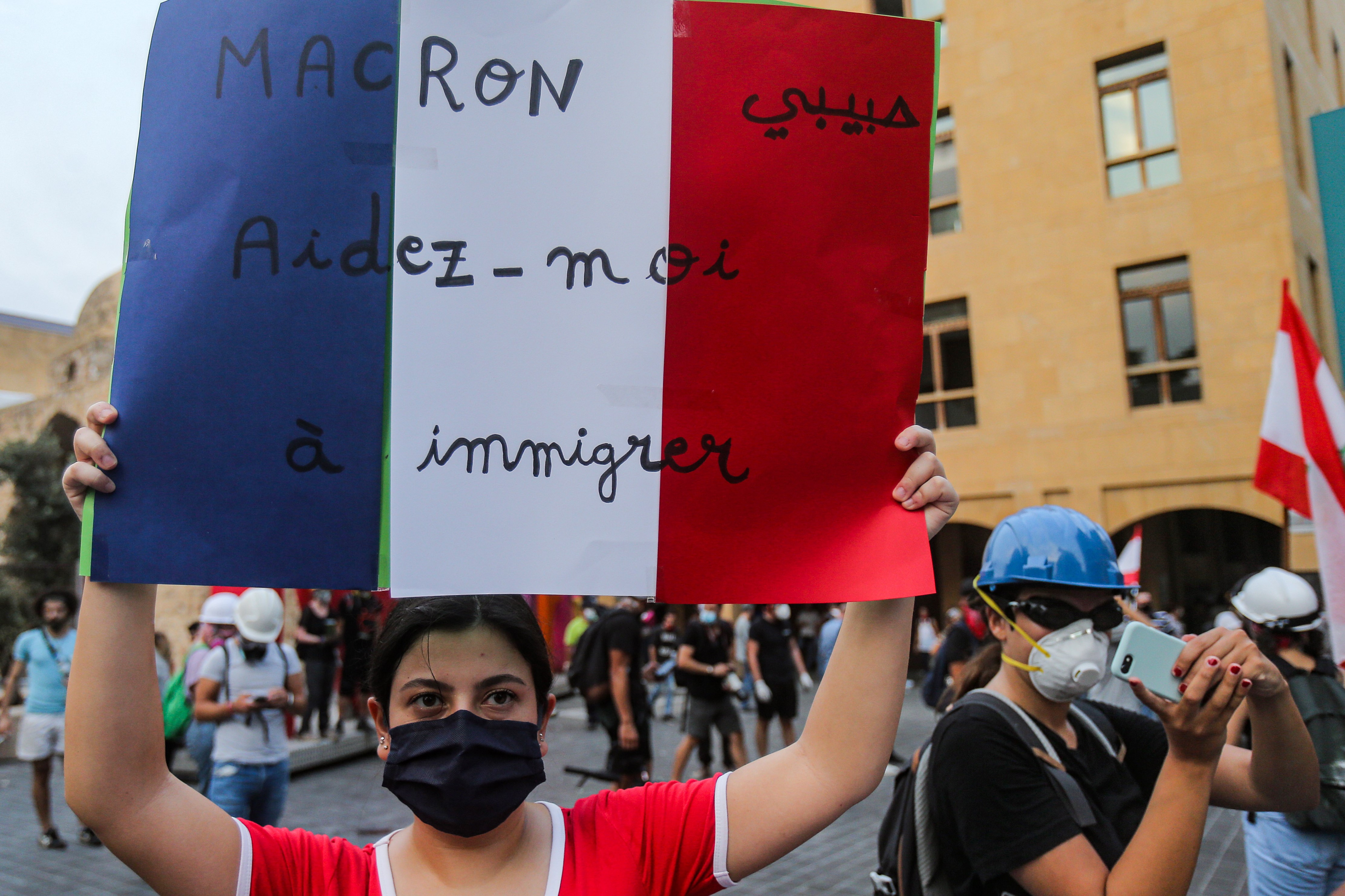 A mask-clad protester holds up a sign showing the French flag with text in Arabic and French reading "Macron, my love, help me to immigrate"