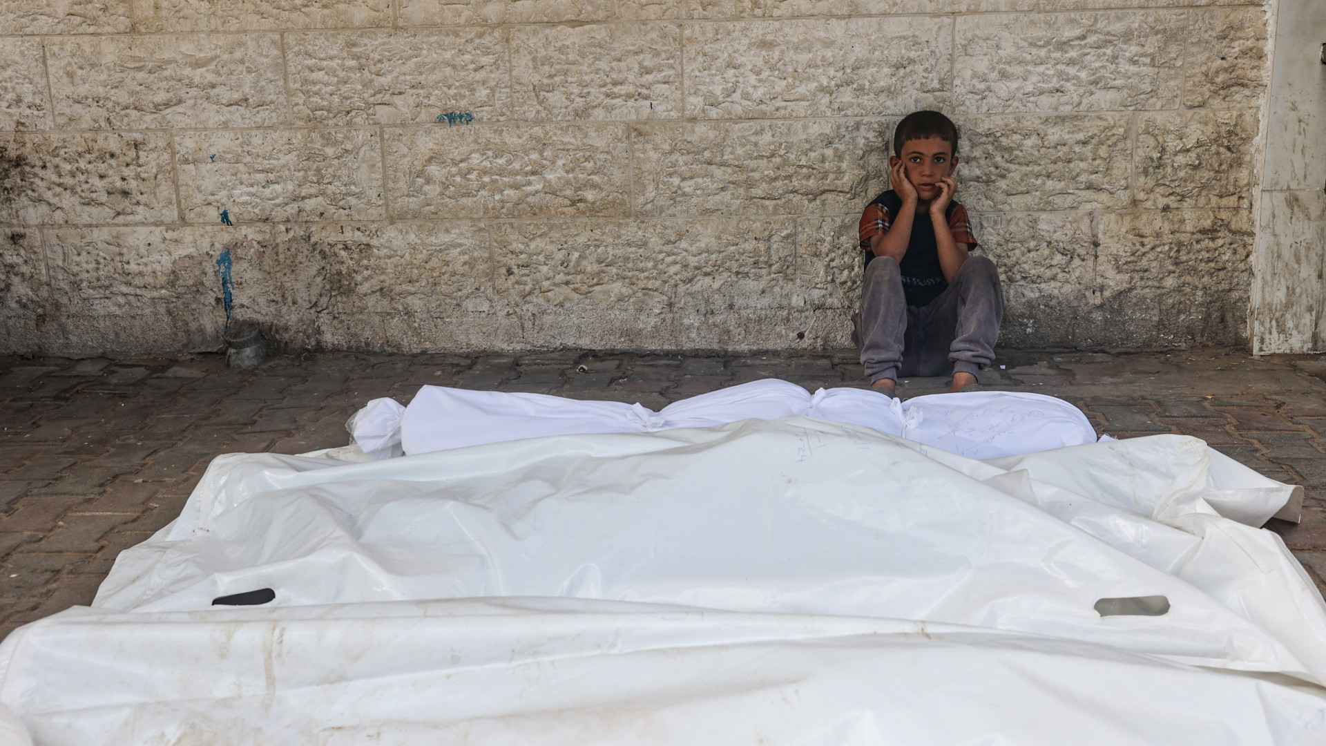 A child sits beside the shrouded corpses of people killed in an overnight Israeli strike, in the yard of the Al-Aqsa Martyrs hospital in Deir el-Balah in central Gaza on 10 August (Eyad Baba/AFP)