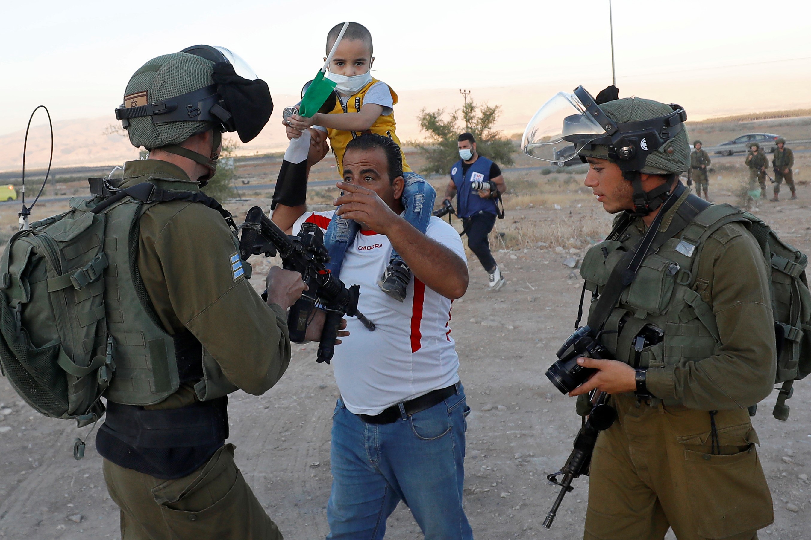 A Palestinian man carrying a chid as the Israeli soldier points the gun at him during a protest against Israel's plan to annex parts of the occupied West Bank, in Jordan Valley on 24  June (Reuters)