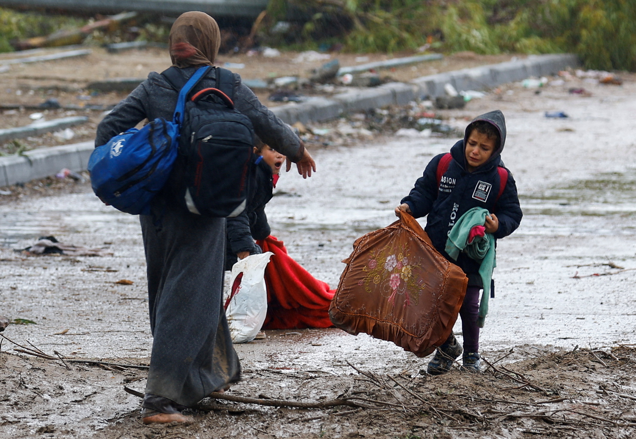 A Palestinian woman fleeing north Gaza helps her children pick up their belongings as they move southward following rainfall on 27 November 2023 (Reuters/Ibraheem Abu Mustafa)