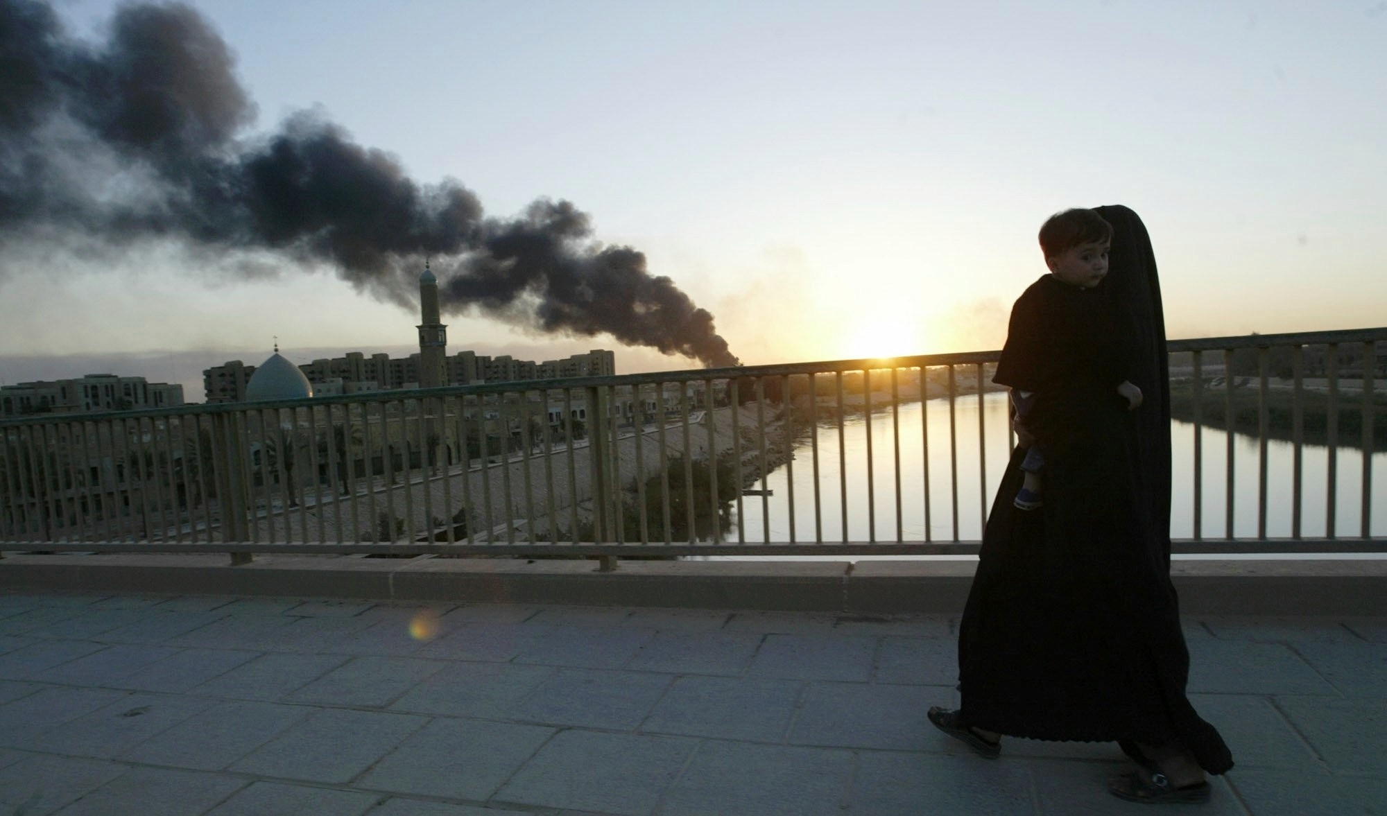 With the fire from a burning warehouse set ablaze by looters in the distance, an Iraqi woman and her child cross the Tigris river, 11 April 2003 (AP)