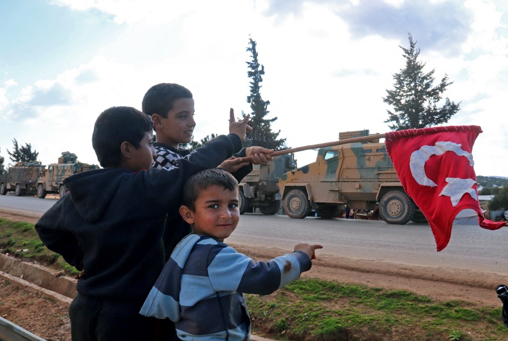 Children wave the Turkish flag as a Turkish military convoy drives near the Syrian town of Batabu on the highway linking Idlib to the Bab al-Hawa border crossing with Turkey, on 2 March 2020 (AFP)