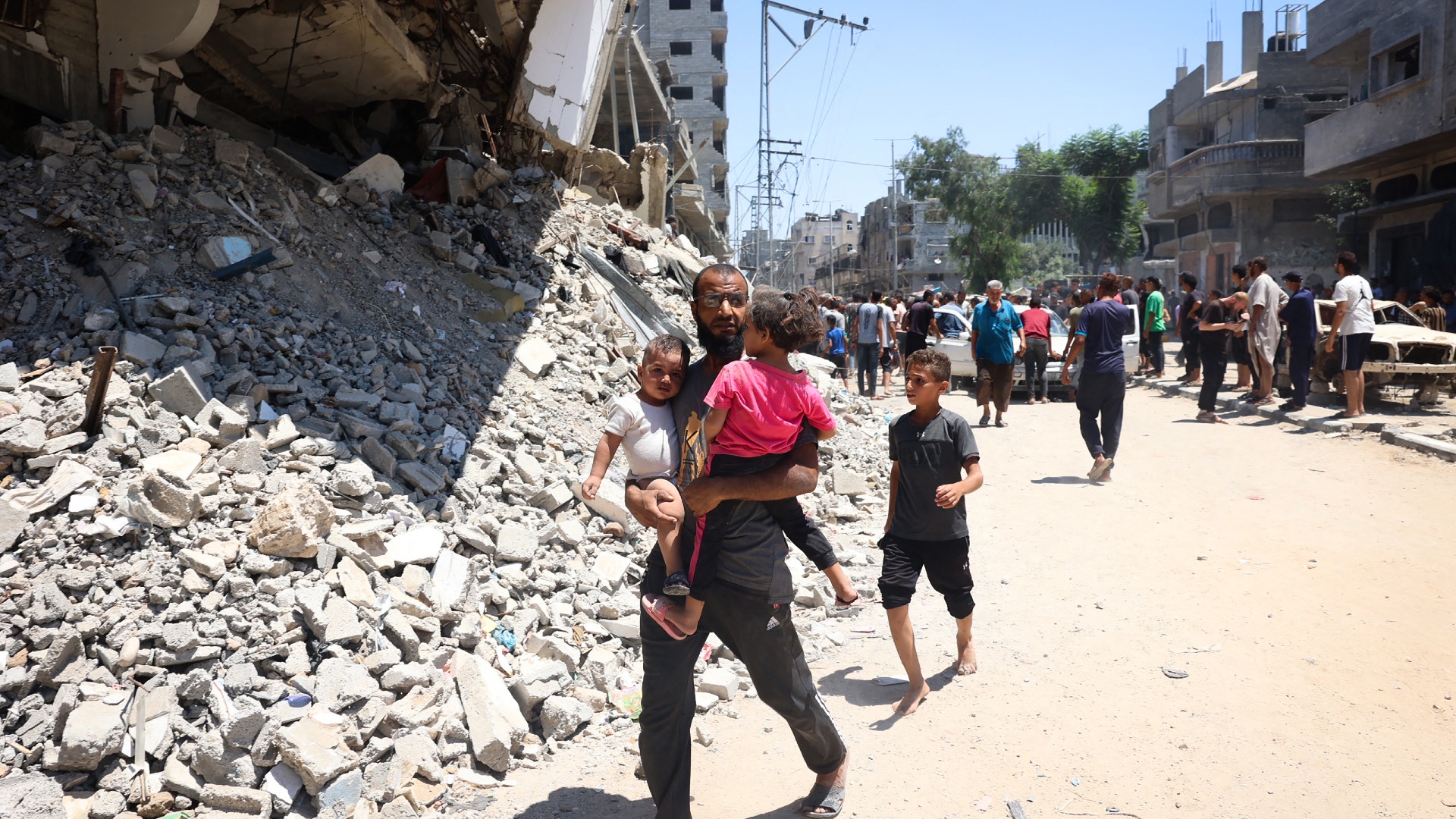 A man carries children as people inspect the damage following Israeli bombardment at al-Bureij refugee camp in the central Gaza Strip on 23 July (Eyad Baba/AFP)