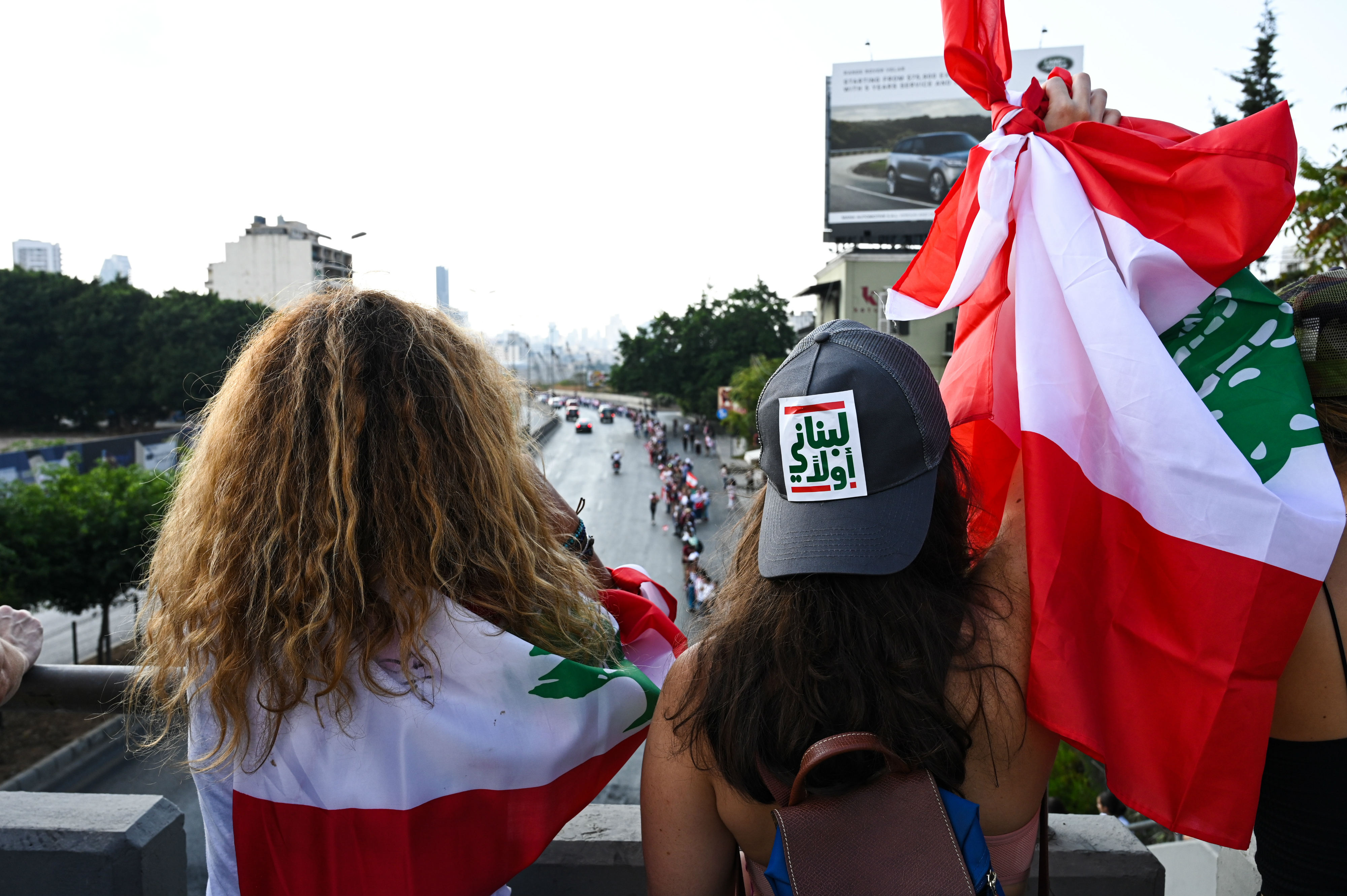 A woman, wearing a hat that reads “Lebanese first”, raises a Lebanese flag as she looks down on the human chain from a bridge above the highway (MEE/Finbar Anderson)