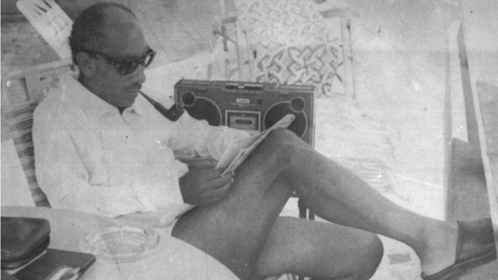 The former Egyptian president Anwar Sadat sits wearing sunglasses, a shirt and shorts while reading a newspaper and listening to a cassette player.