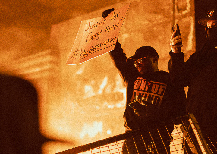 A man holds a sign reading "Justice for George Floyd" in front of a burning building.