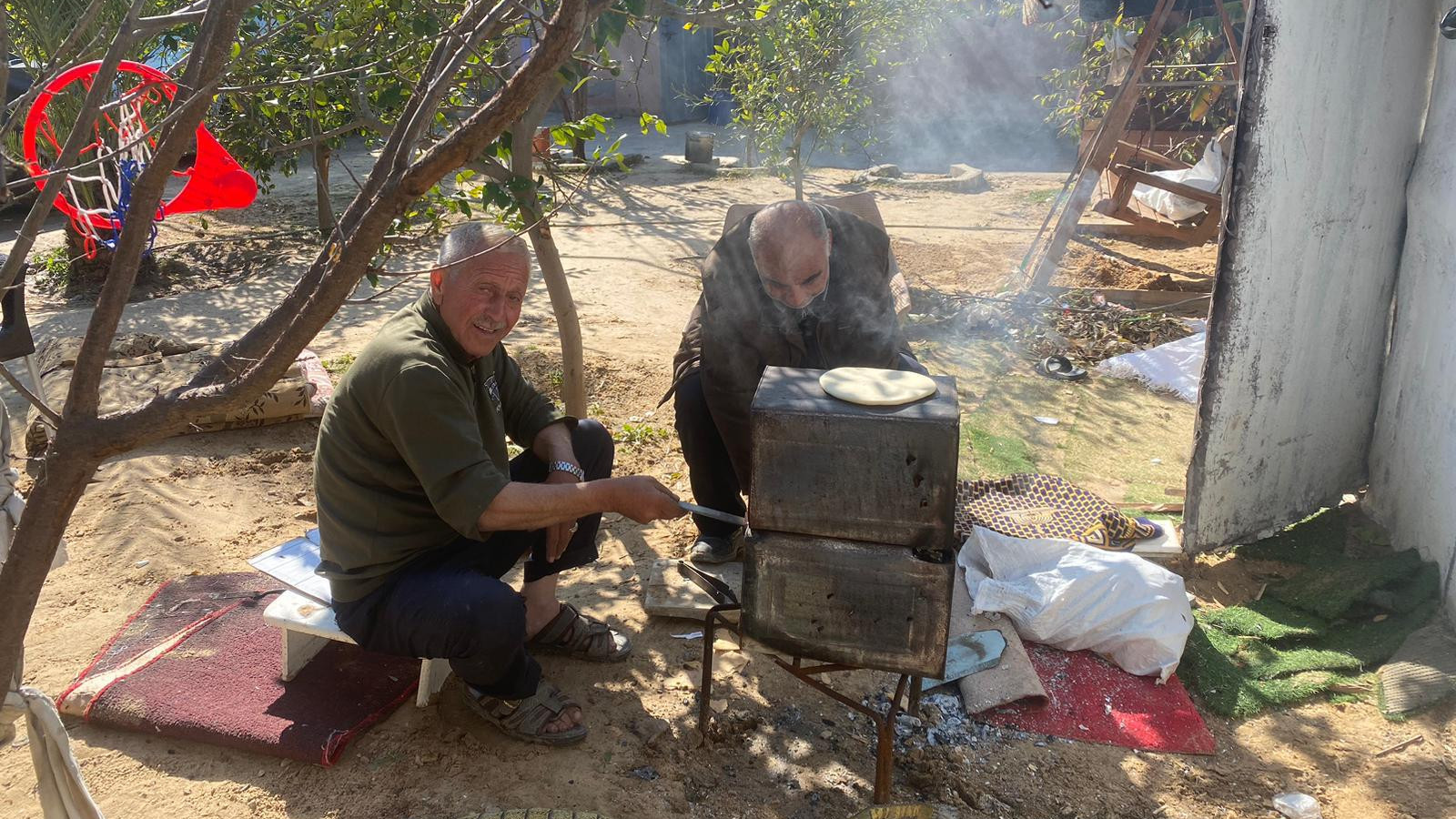 The author's father, left, cooks using a makeshift oven with a relative near the Rafah refugee tent camp in southern Gaza after being forcibly displaced in early May (Supplied)