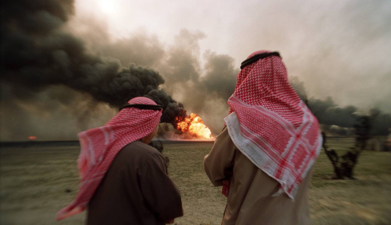 Two Kuwaiti men watch in Al Ahmadi oil field watch a burning oil well set ablaze by Iraqi troops on 26 March 1991 (AFP)