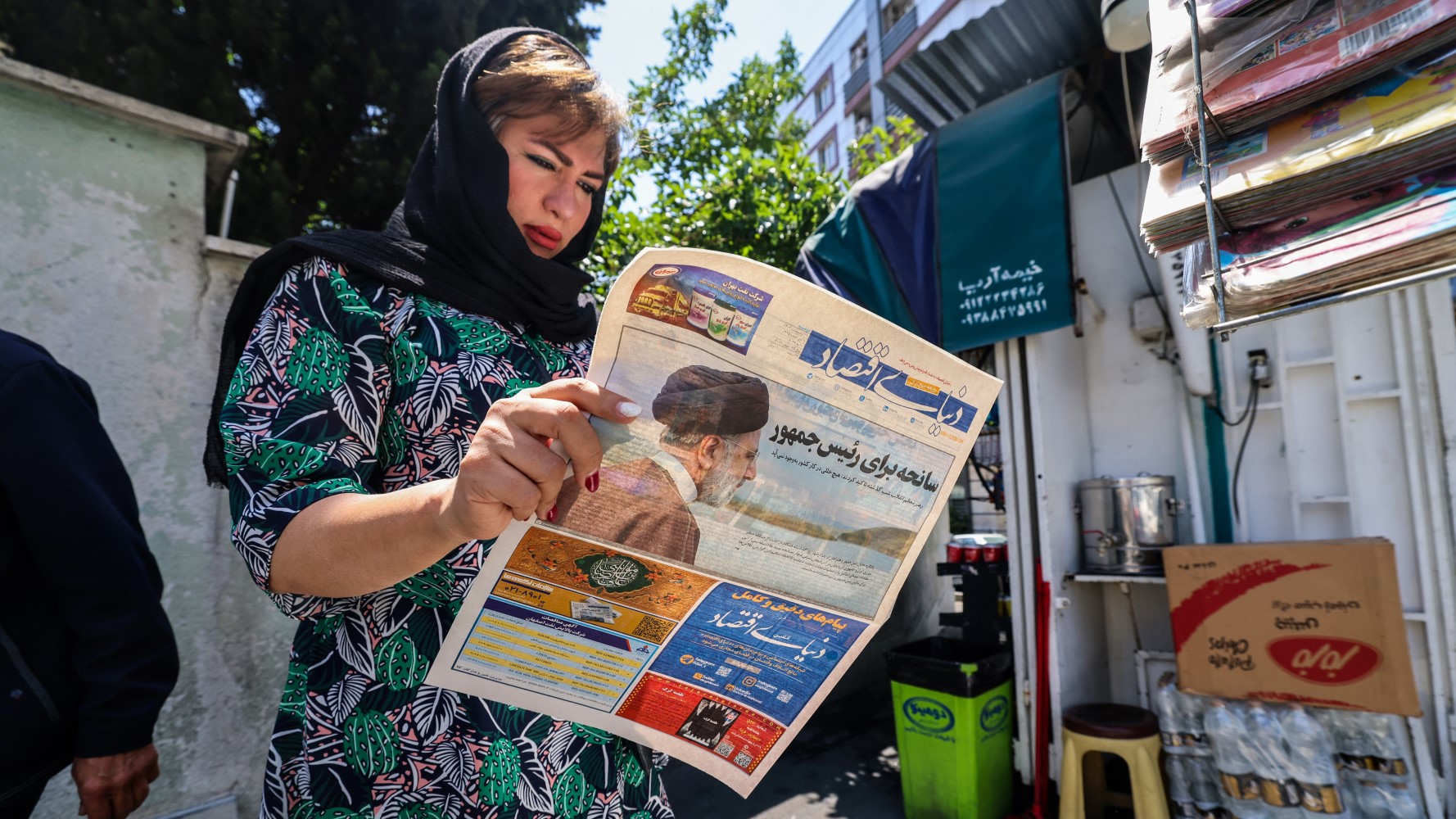 A woman reads a newspaper with a front-page report on the crash of the Iranian president's helicopter outside a kiosk in Tehran on May 20, 2024. Iranian President Ebrahim Raisi was declared dead on May 20 after rescue teams found his crashed helicopter in a fog-shrouded western mountain region, sparking mourning in the Islamic republic. Atta KENARE 