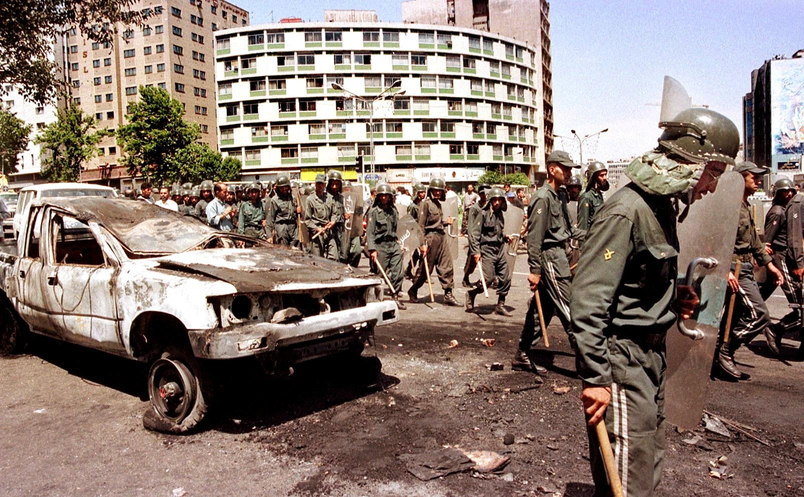 Iranian anti-riot police pass by a destroyed car in the centre of Tehran on 12 July 1999 (AFP)