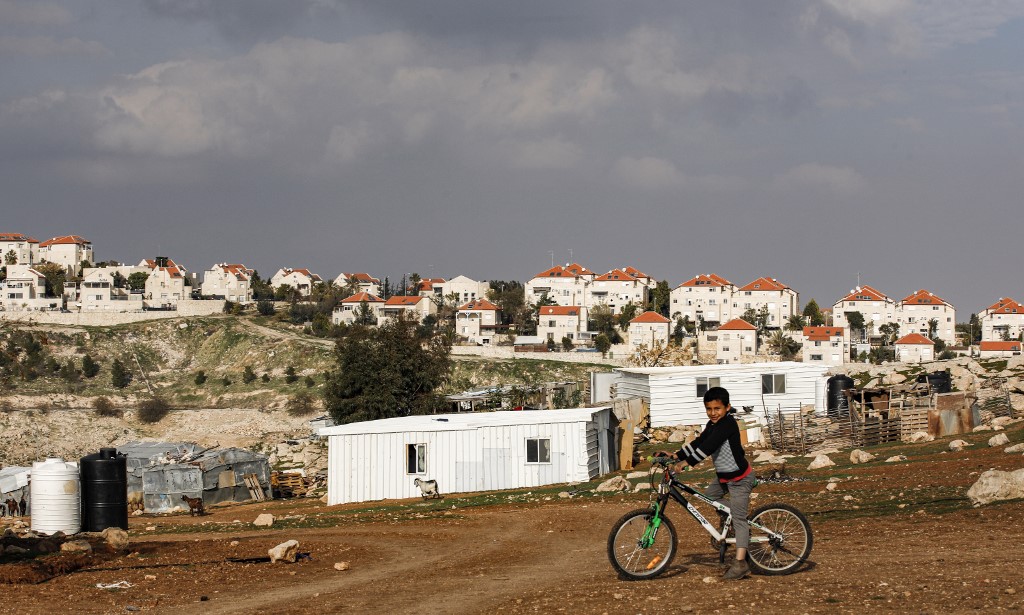 A boy rides a bicycle past Palestinian Bedouin huts in the occupied West Bank on 28 January, with the Israeli settlement of Maale Adumim in the background (AFP)