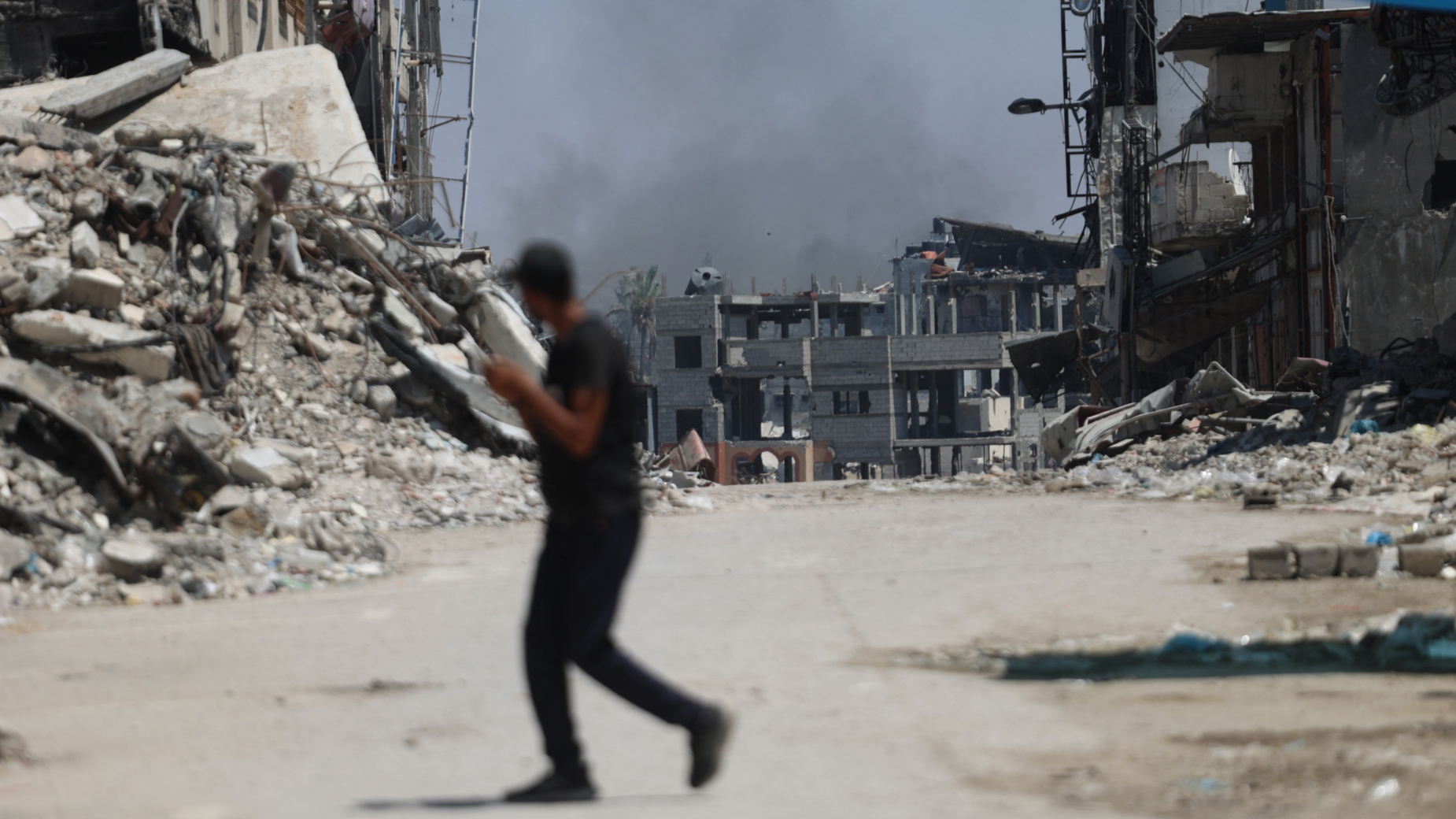 A Palestinian man crosses a street as smoke billows following an Israeli strike on Gaza City's Shujaiya on 28 June 2024 (Omar al-Qattaa/AFP)