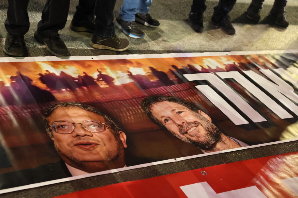 Israelis stand next to a poster bearing the portraits of Itamar Ben-Gvir and Bezalel Smotrich during a protest against the government in Tel Aviv on 4 March 2023 (AFP)