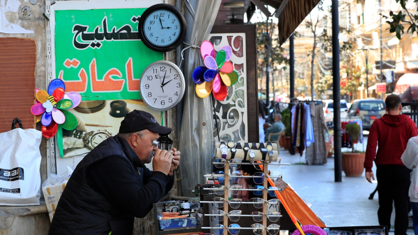 A Lebanese street vendor sits next to two clocks that show different times in Lebanon, in the southern port city of Sidon, 27 March 2023 (AP)