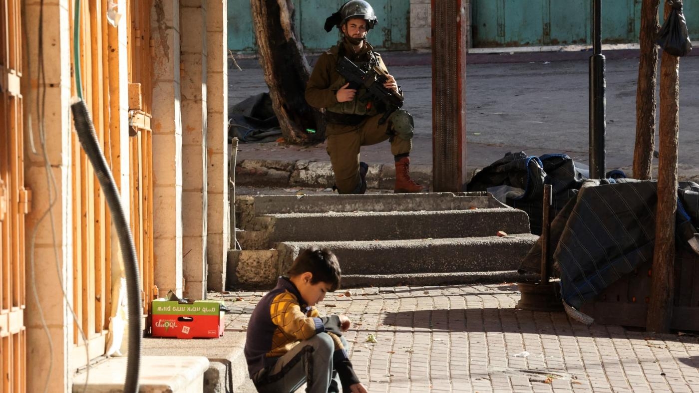 Un jeune palestinien assis sur les marches d’un magasin fermé alors qu’un membre des forces de sécurité israéliennes prend position à Hébron, le 21 janvier 2022 (AFP)