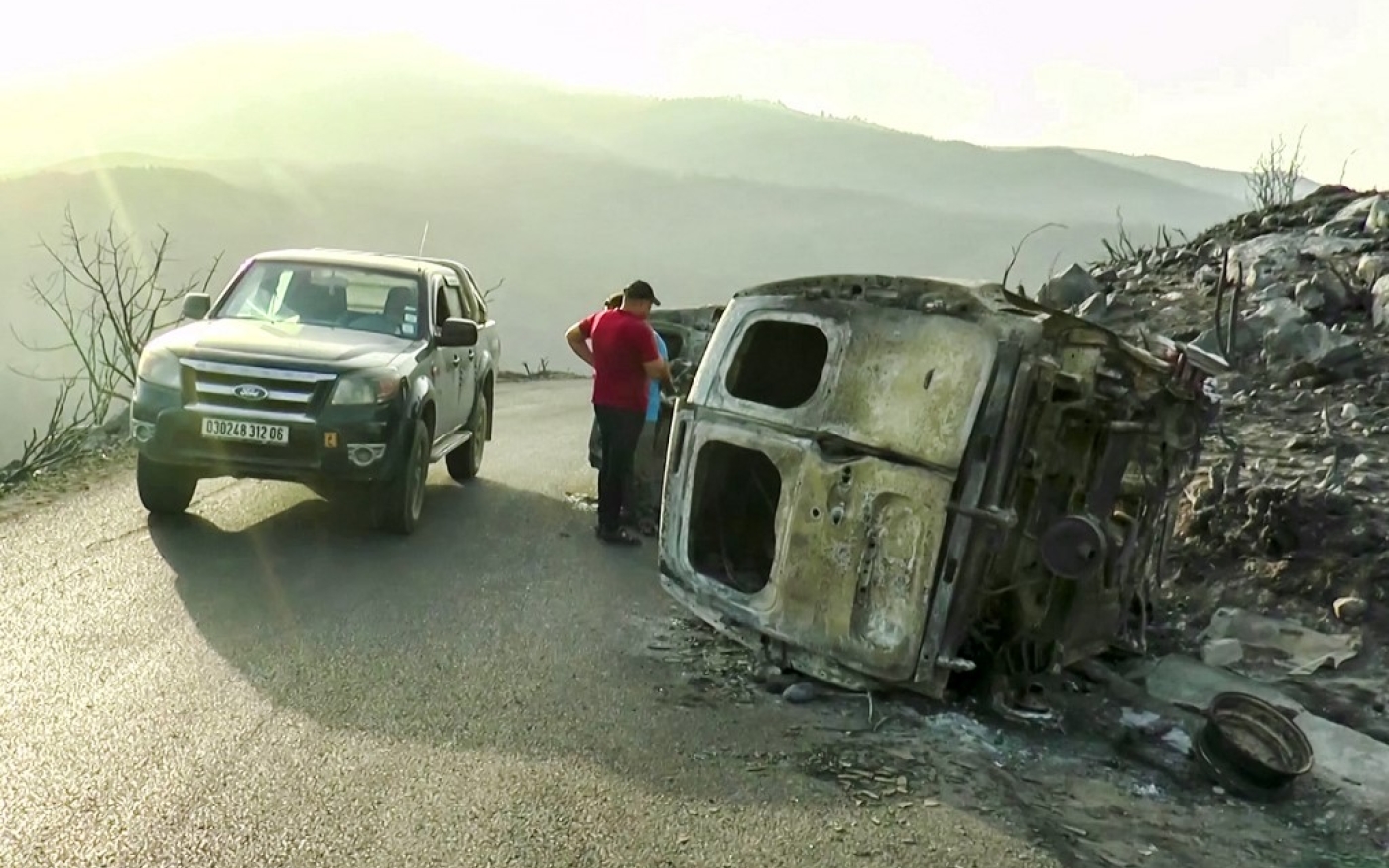 Des personnes inspectent des véhicules incendiés sur le bord d’une route de montagne à la suite d’incendies dans les forêts de Béjaïa, au nord-est d’Alger, le 25 juillet 2023 (AFP)