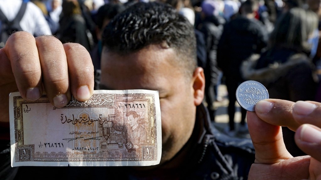 A Syrian man holds a Syrian lira note and a coin covering his face while standing in a busy shopping festival in Damascus.
