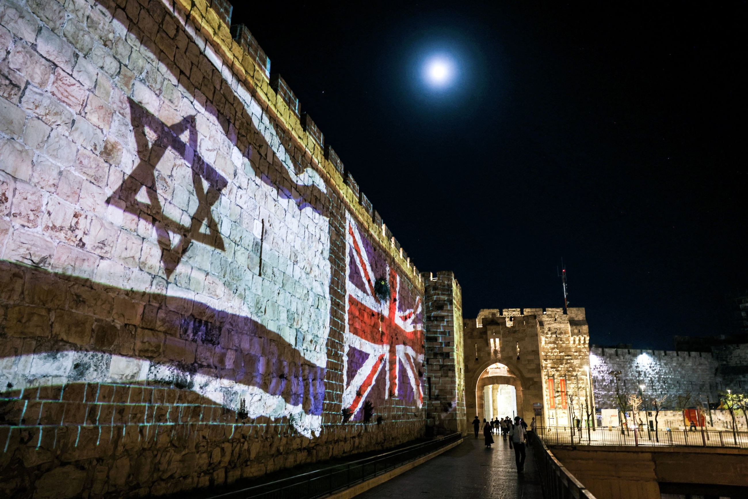 People walk past projections of the flags of Israel and the British Union Jack displayed on the walls of the old city of Jerusalem on September 10, 2022, days following the death of Queen Elizabeth II of the United Kingdom (AFP)