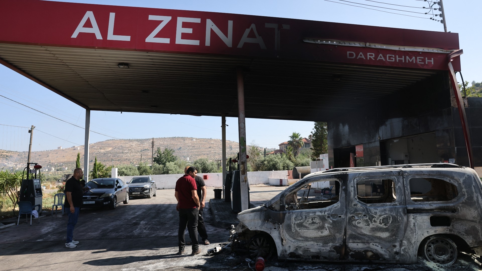 People stand near a burnt car, reportedly set ablaze by Israeli settlers, at a petrol station in Lubban al-Sharqiya in the occupied West Bank on 21 June (AFP)