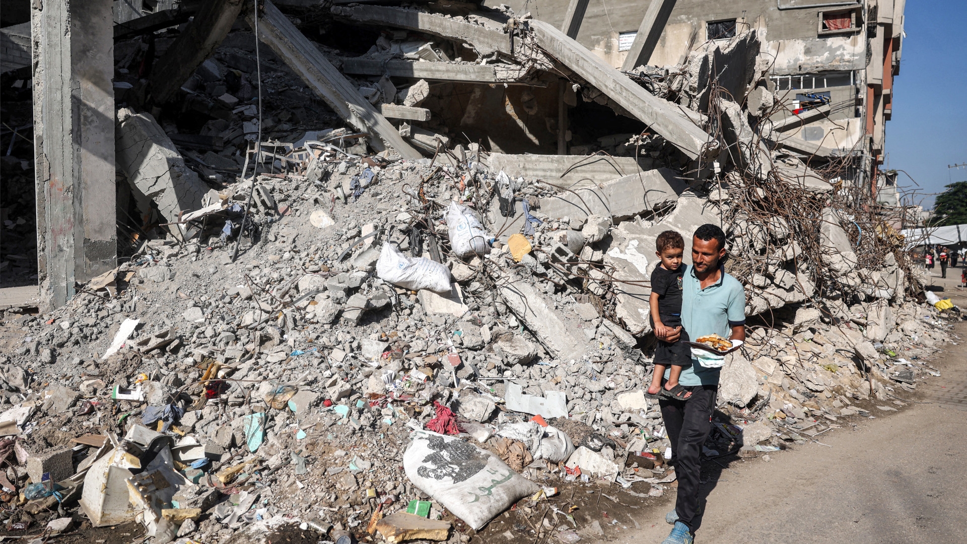 A man carries a child while walking past a building destroyed by Israeli bombardment in the Bureij camp for Palestinian refugees in the central Gaza Strip on 9 October 2024 (Eyad Baba/AFP)