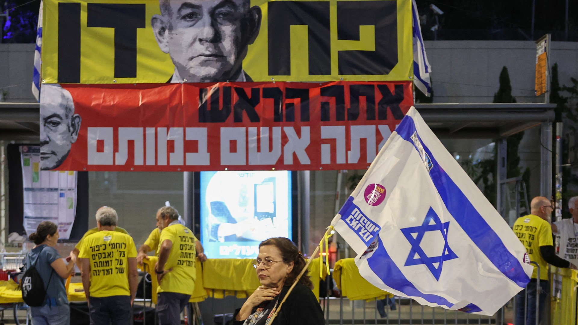 A demonstrator lifts an Israeli flag as she walks past a banner depicting Israeli Prime Minister Benjamin Netanyahu with the word "Fearful" in Hebrew during an anti-government protest demanding action for the release of Israelis held captive in Gaza in front of the Defence Ministry in Tel Aviv on 23 November 2024 (Jack Guez/AFP)