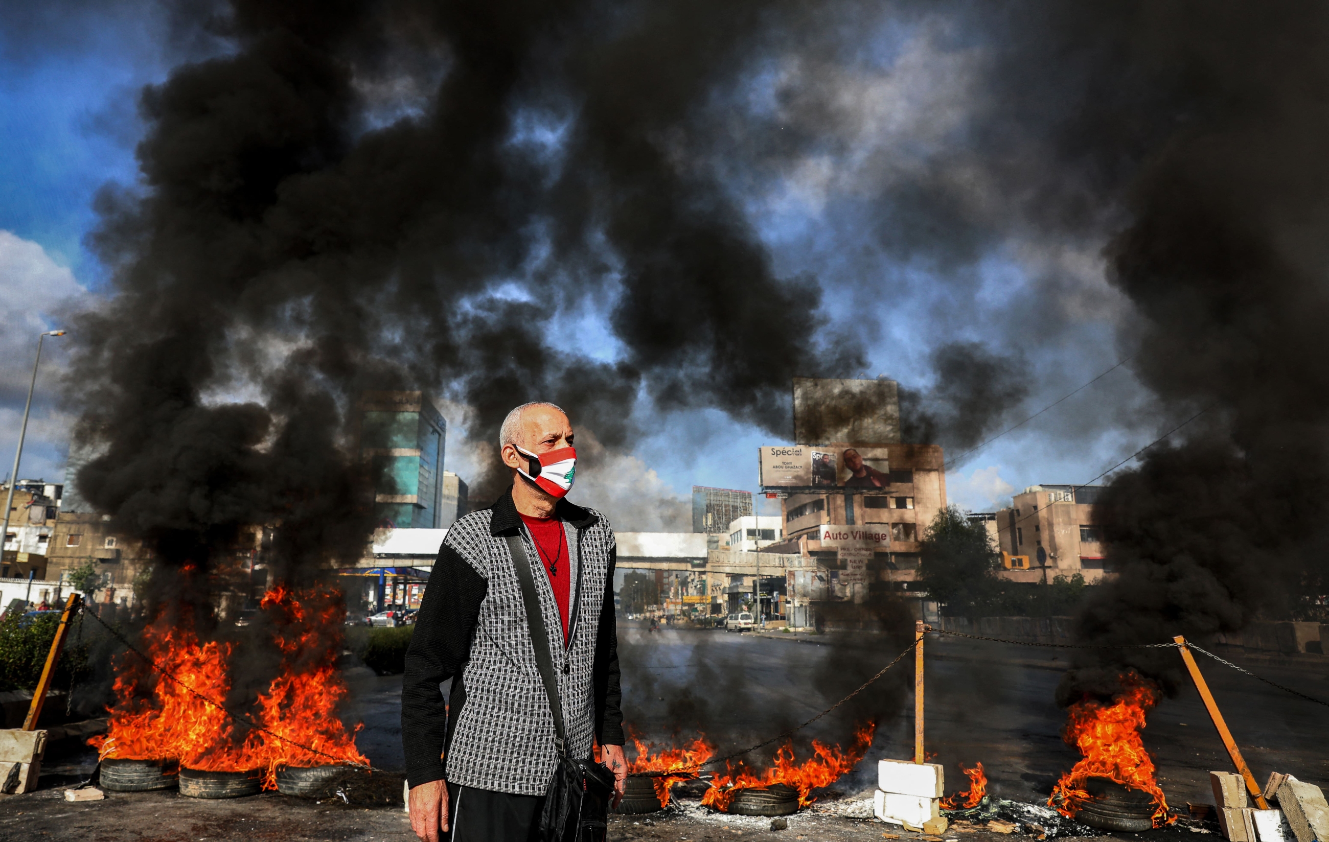A man in mask a depicting the Lebanese flag stands next to flaming tires at a make-shift roadblock set-up by anti-government demonstrators in  Dora on the northern outskirts of Lebanon's capital Beirut on 8 March 8 2021 (Anwar Amro/AFP)