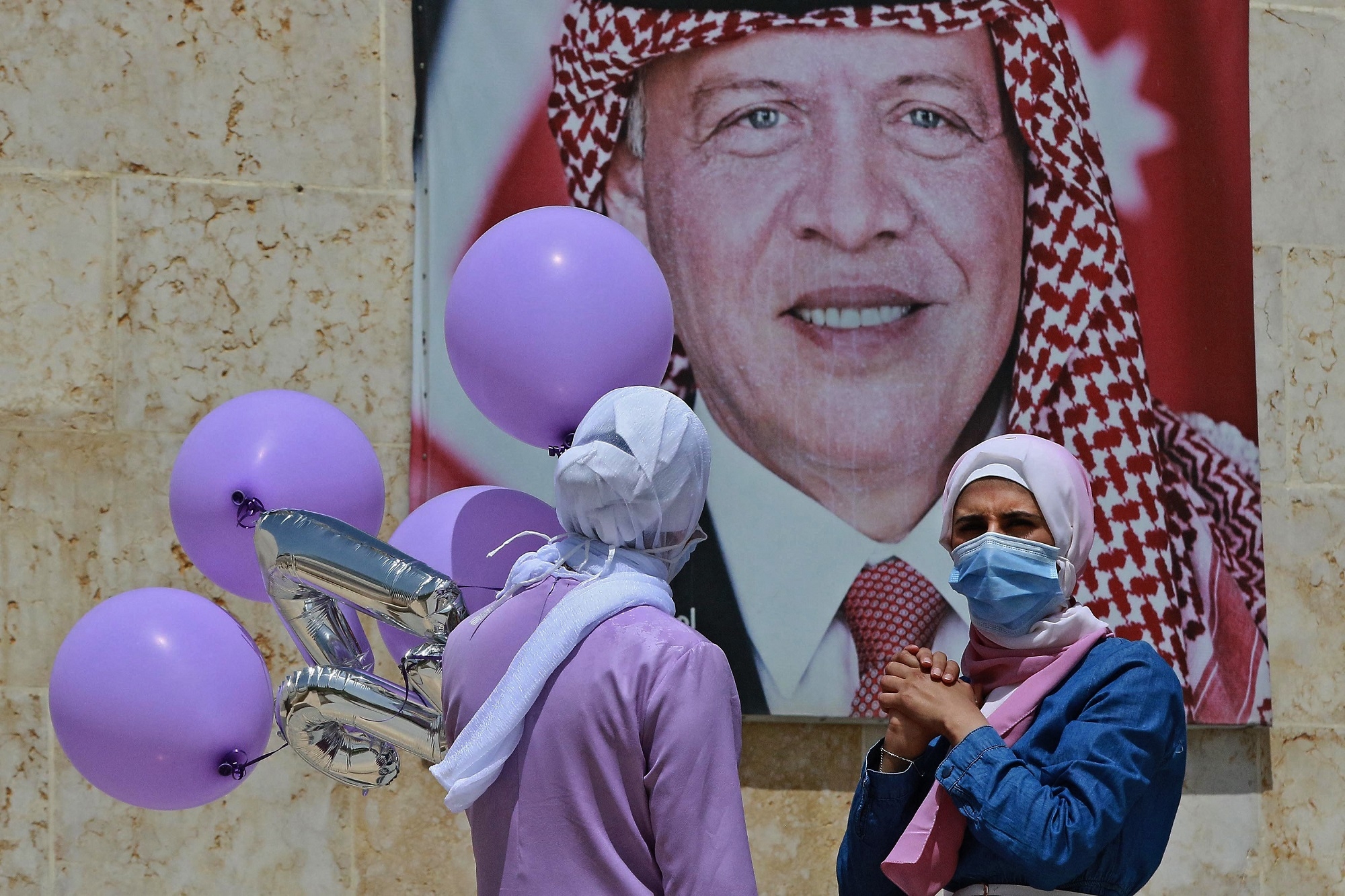 Women carrying balloons stand next to a poster of Jordan's King Abdullah in Amman