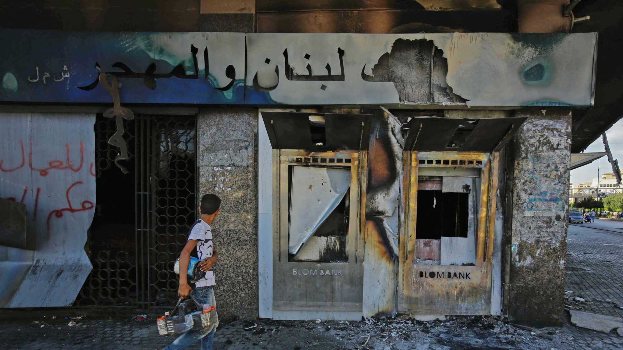 A youth walks past a burnt down branch of Blom Bank after it was set on fire and vandalised by protesters in al-Nour Square in Lebanon's northern city of Tripoli on 12 June 2020 (AFP)