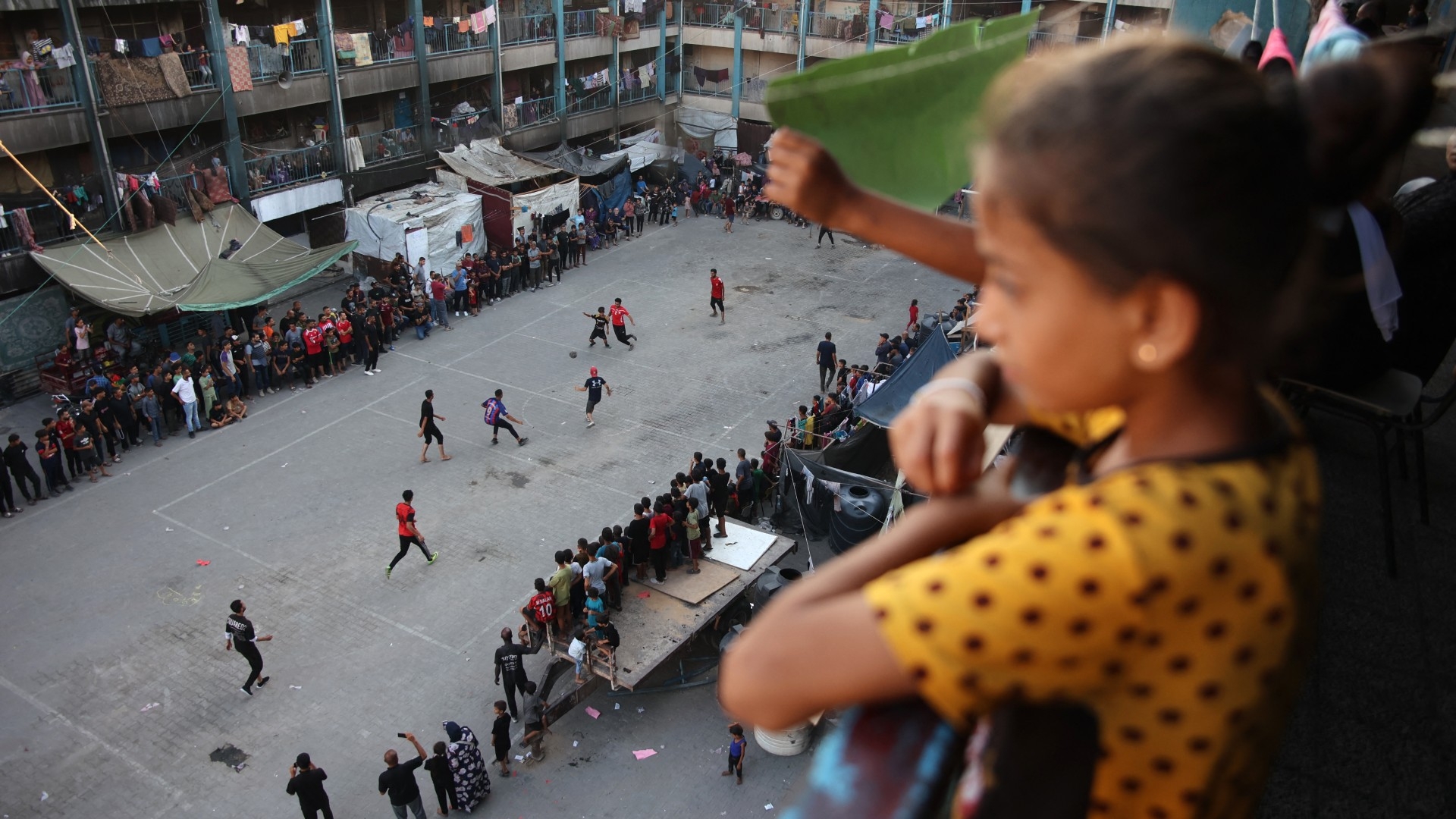 Children watch as displaced Palestinians play football in the courtyard of a UN-run school in Jabalia, in the northern Gaza Strip on 23 July 2024 (AFP/Omar al-Qattaa)