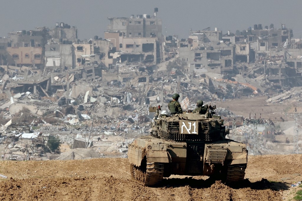 A picture taken from a position in southern Israel along the border with the Gaza Strip shows an Israeli tank rolling along the fence as damaged buildings are see in the Gaza strip on 19 January 2024 (AFP)
