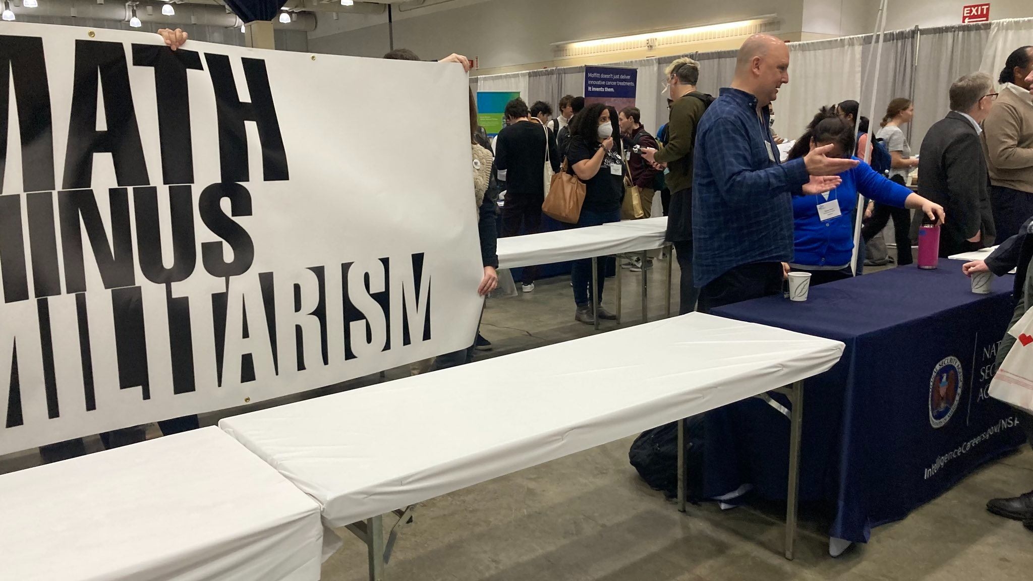Activists hold a banner reading "MATH MINUS MILITARISM" next to a table for the NSA during a job recruitment fair at the Joint Mathematics Meeting on 5 January 2023.