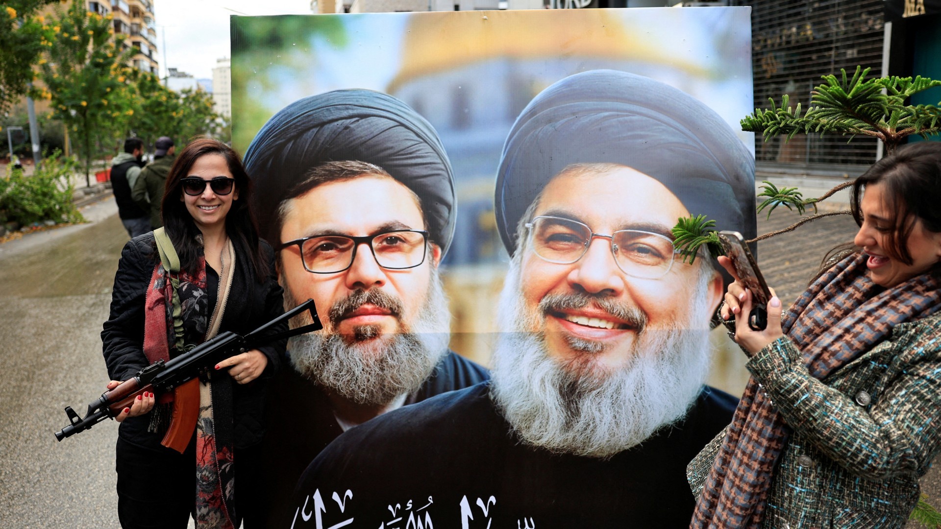 A woman holding a weapon stands next to a poster with the images of Hassan Nasrallah, right, and Hashem Safieddine, at the entrance of Beirut's southern suburbs on 27 November 2024 (Reuters/Thaier Al-Sudani)