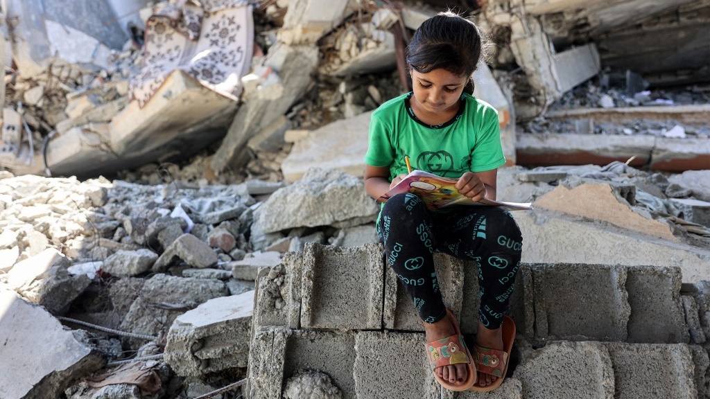 A girl writes in a notebook by the rubble of destroyed buildings in Jabalia, Gaza, on 8 September 2024 (Omar al-Qattaa/AFP)