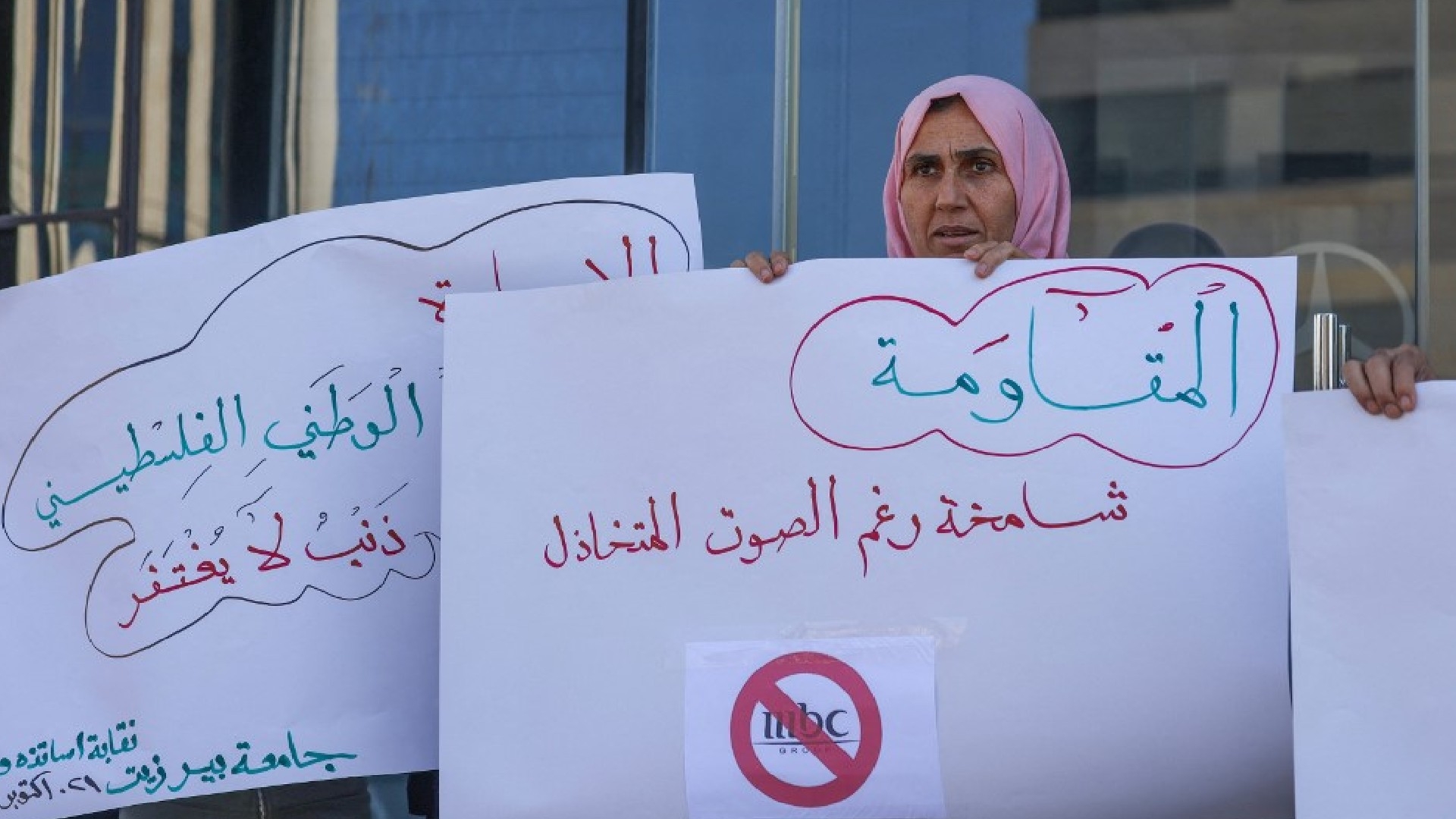 Palestinians lift placards during a rally at the entrance of a building housing the offices of Saudi broadcasters MBC, Al-Arabiya, and Al-Hadath in Ramallah in the occupied West Bank on 21 October 2024 (AFP/Jaafar Ashtiyeh)