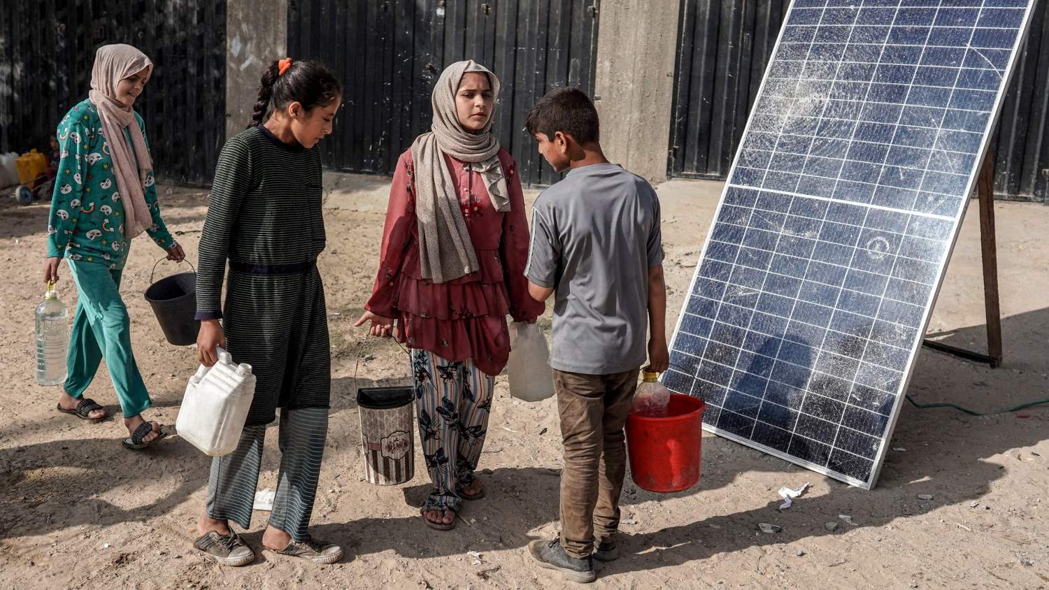 Palestinian children walk past a solar panel while carrying jerrycans and pails of water as people collect water from a tanker cistern in Deir el-Balah in the central Gaza Strip on 30 April 2024.