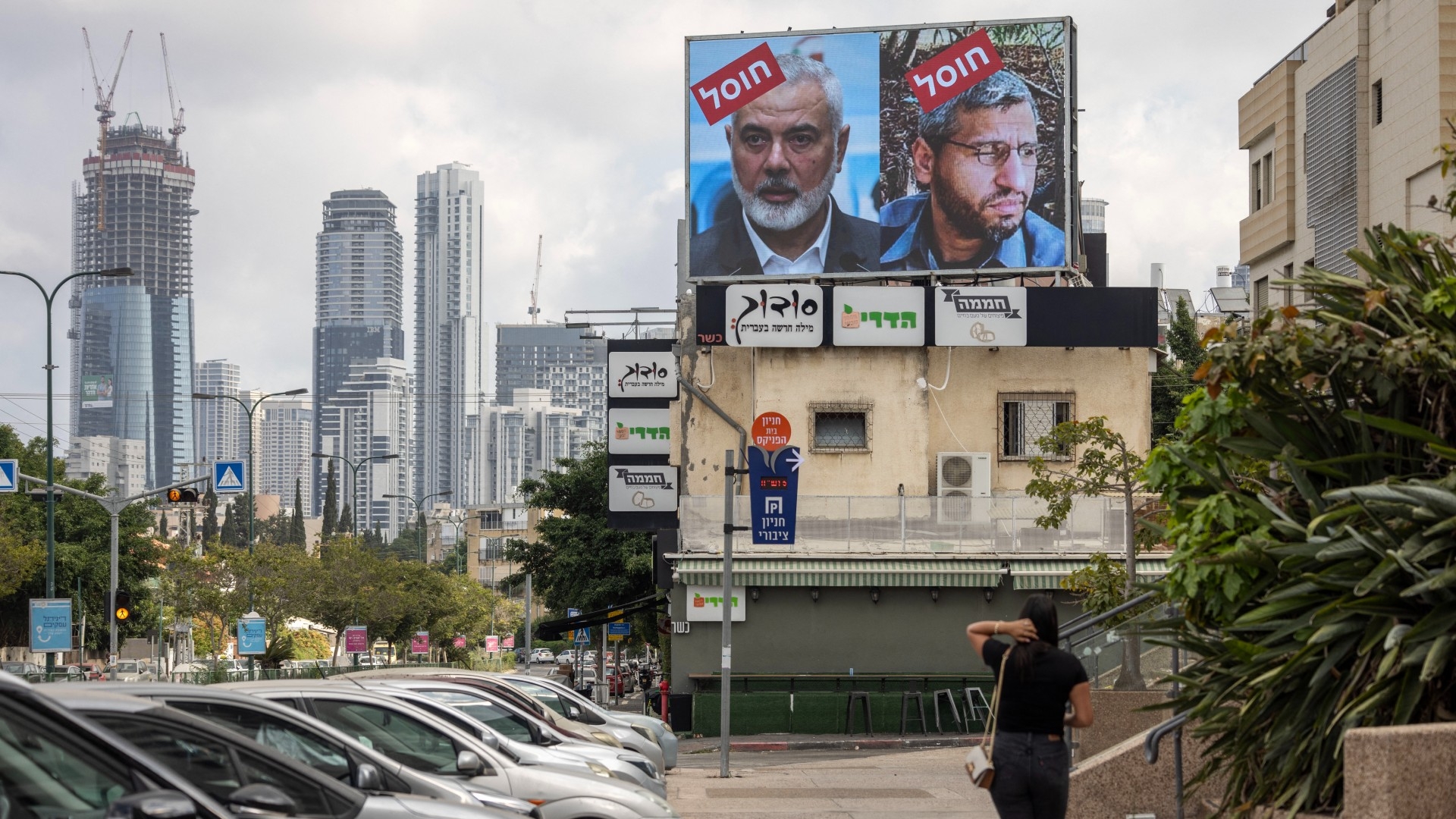 A woman walks towards a billboard displaying portraits of Hamas leaders Mohammed Deif and Ismail Haniyeh with the word “assassinated” written in Hebrew in Tel Aviv on 2 August 2024 (AFP/Oren Ziv)