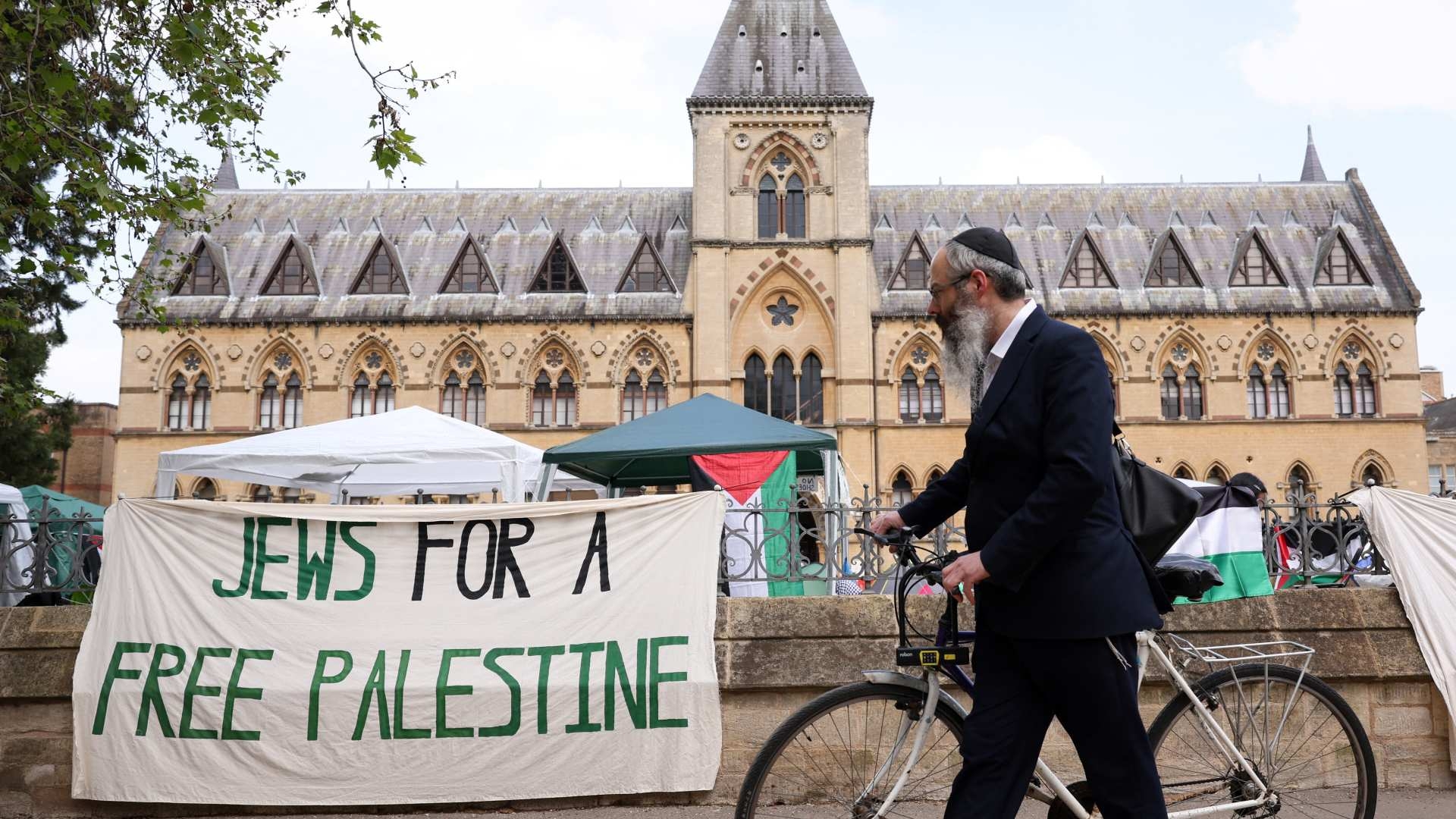 A passerby wearing a kippah pushes his bicycle past an encampment protesting against the war in Gaza at Oxford University on 7 May 2024 (AFP)