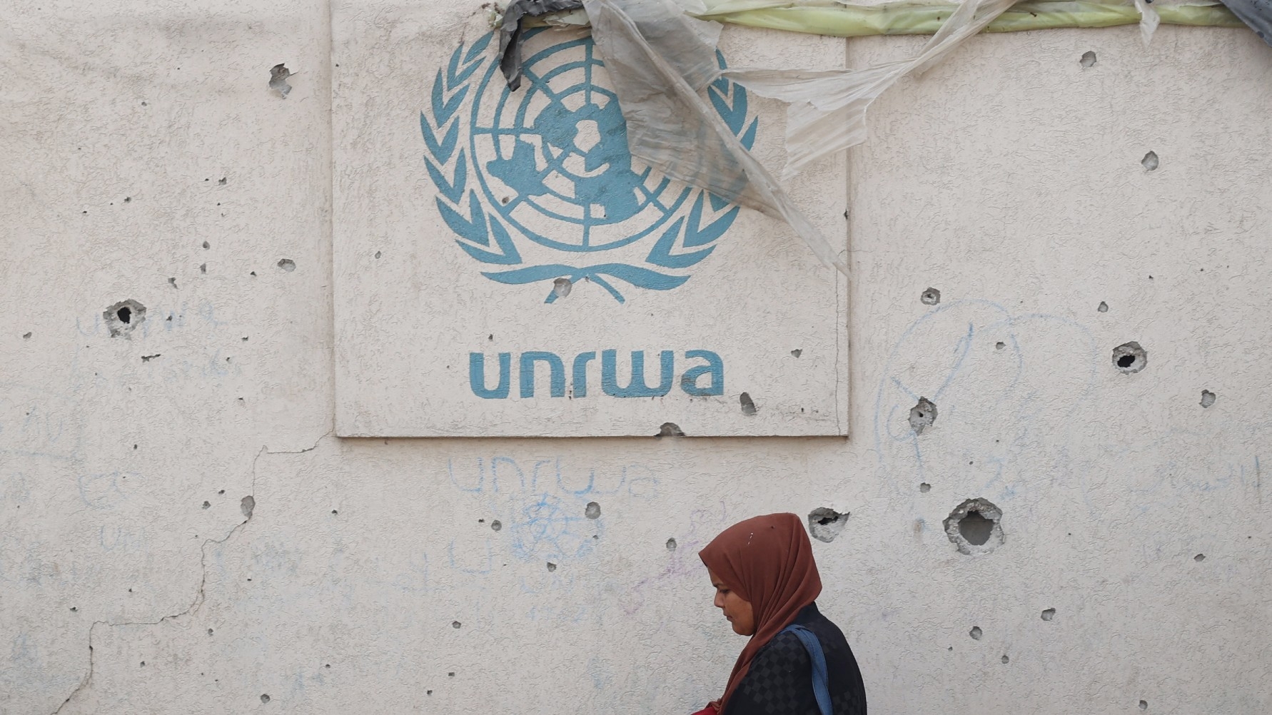 A Palestinian woman walks past a damaged wall bearing the Unrwa logo at a camp for internally displaced people in Rafah in the southern Gaza Strip on 28 May 2024 (AFP)