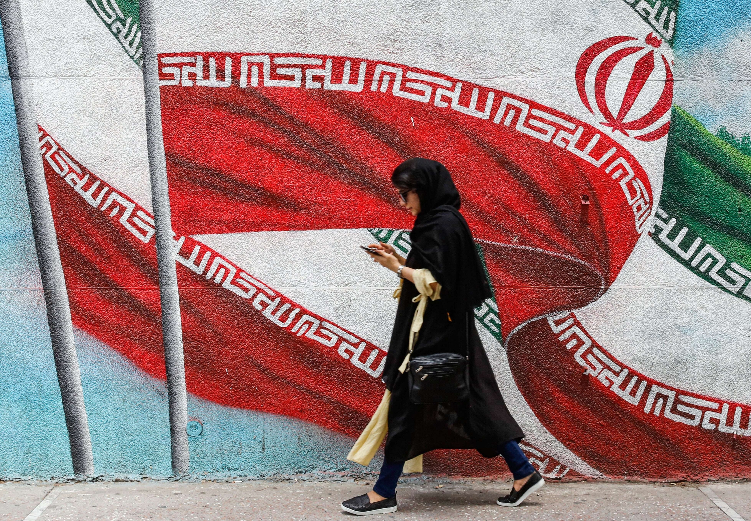 An Iranian girl walks next to a wall painting in Tehran of Iran's national flag while using her phone