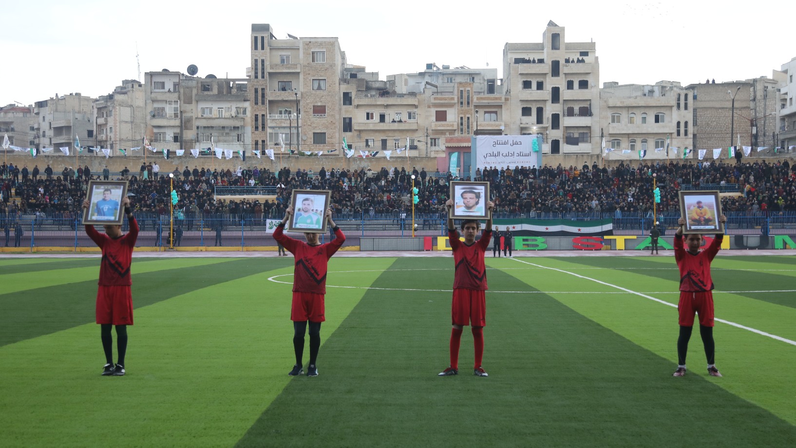Syrian children raise pictures of slain Syria on a pitch in Idlib during the opening ceremony of the 'Camps World Cup' (MEE/Ali Haj Suleiman)
